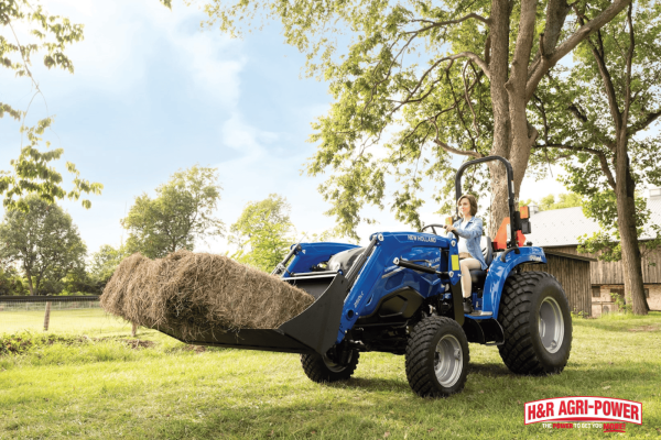 New Holland compact tractor handling hay near a barn and trees, representing proper storage and environmental protection for equipment longevity