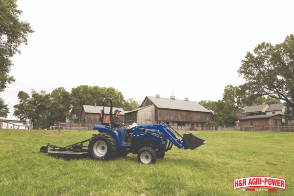 New Holland compact tractor mowing grass near a barn, representing warranty coverage and support services for farm equipment